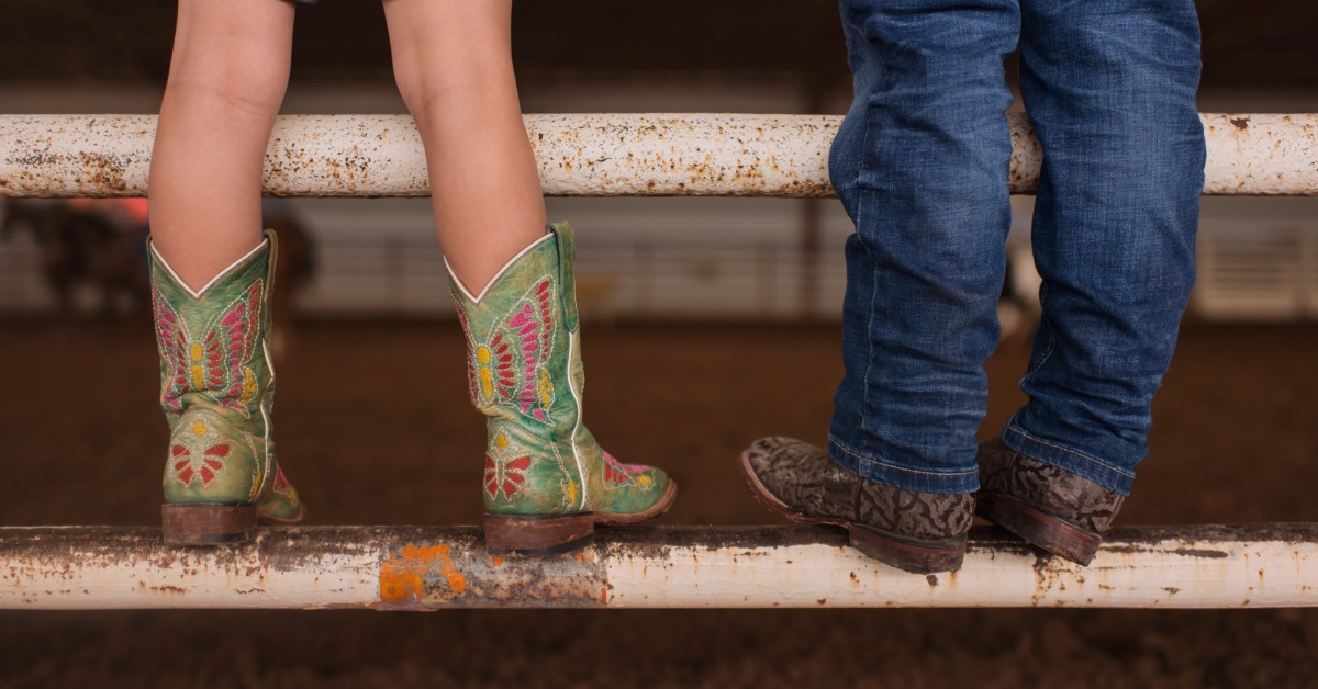 Two children wearing western boots and denim, standing on white metal rails above a dirt floor with a dark background.