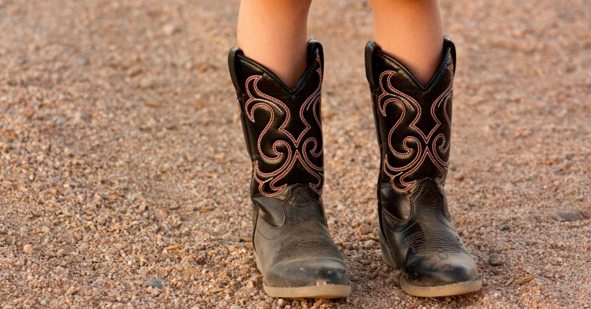 A child wearing black western boots with red stitching, standing on a gravel surface in dark shorts and bare legs.