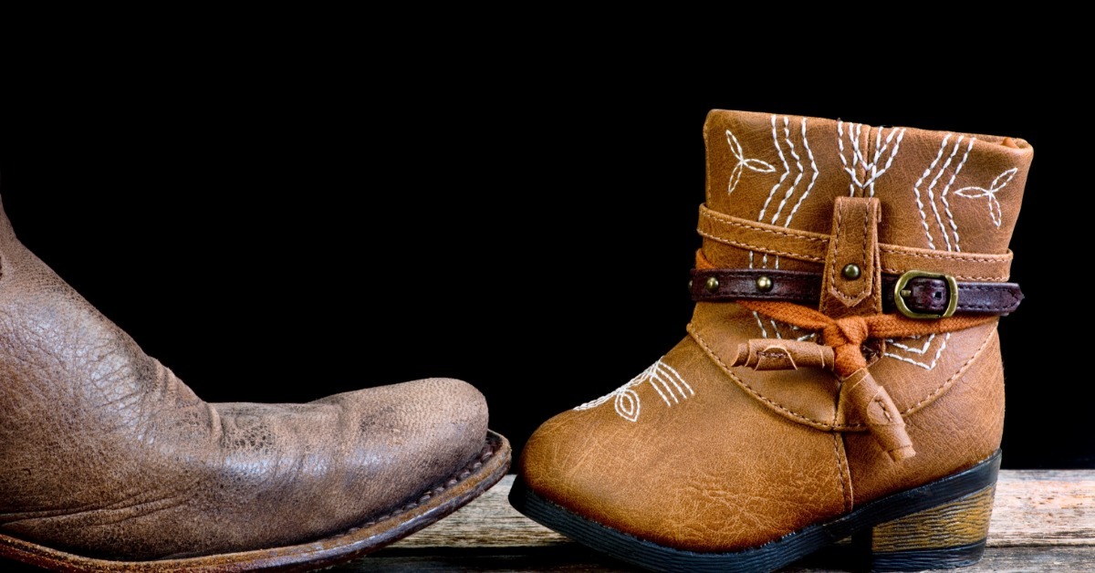 A large worn boot and a small brown western boot sitting face to face on wood against a plain black background.