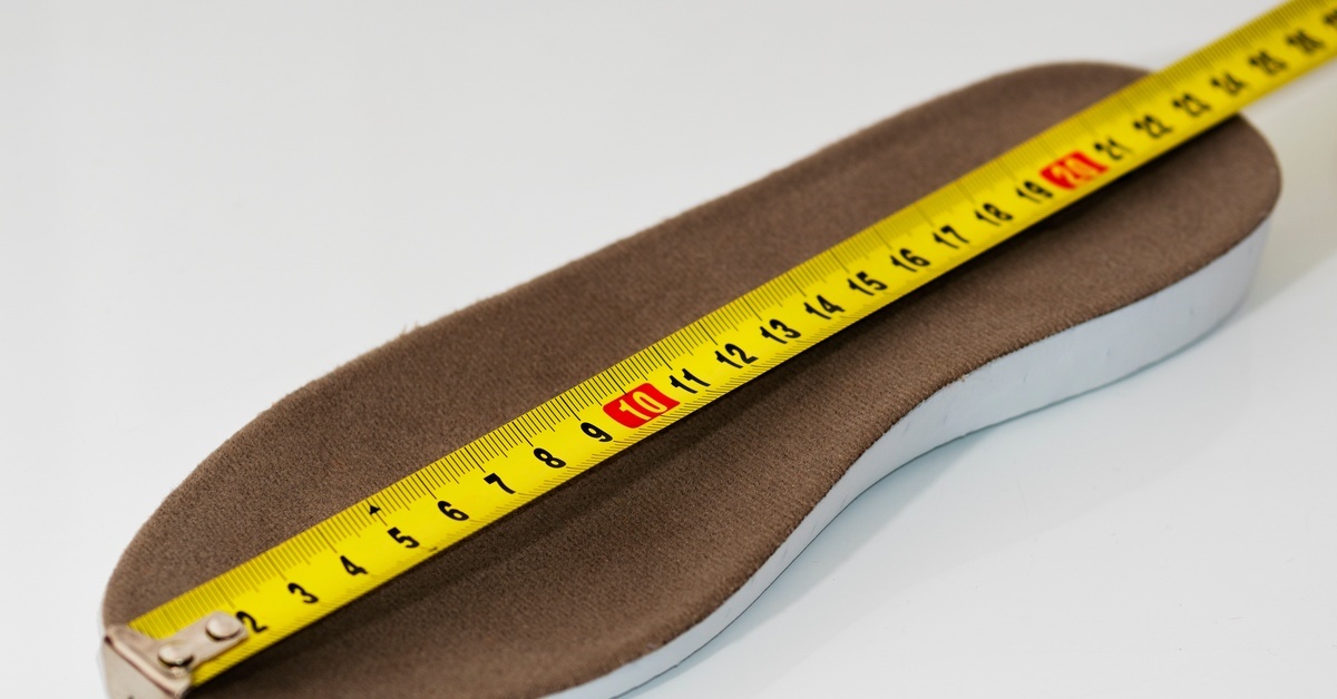 A yellow measuring tape stretching across a brown shoe insole on a plain white surface with black and red numbers visible.