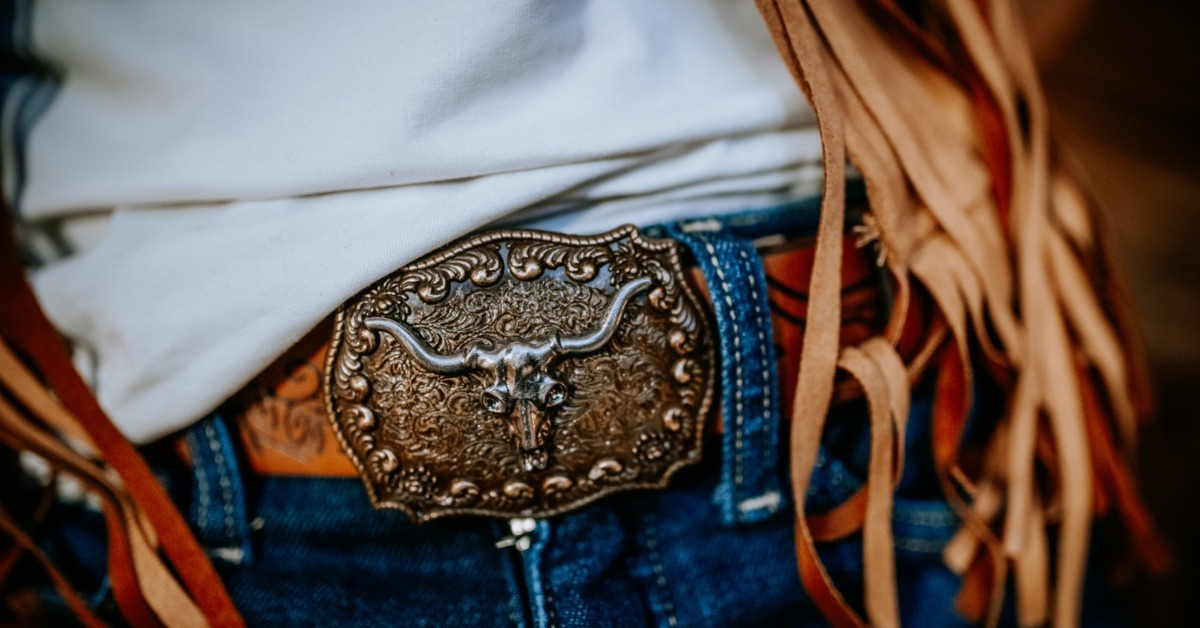 An ornate metal belt buckle with a longhorn skull design worn over blue jeans beside a brown fringed garment.