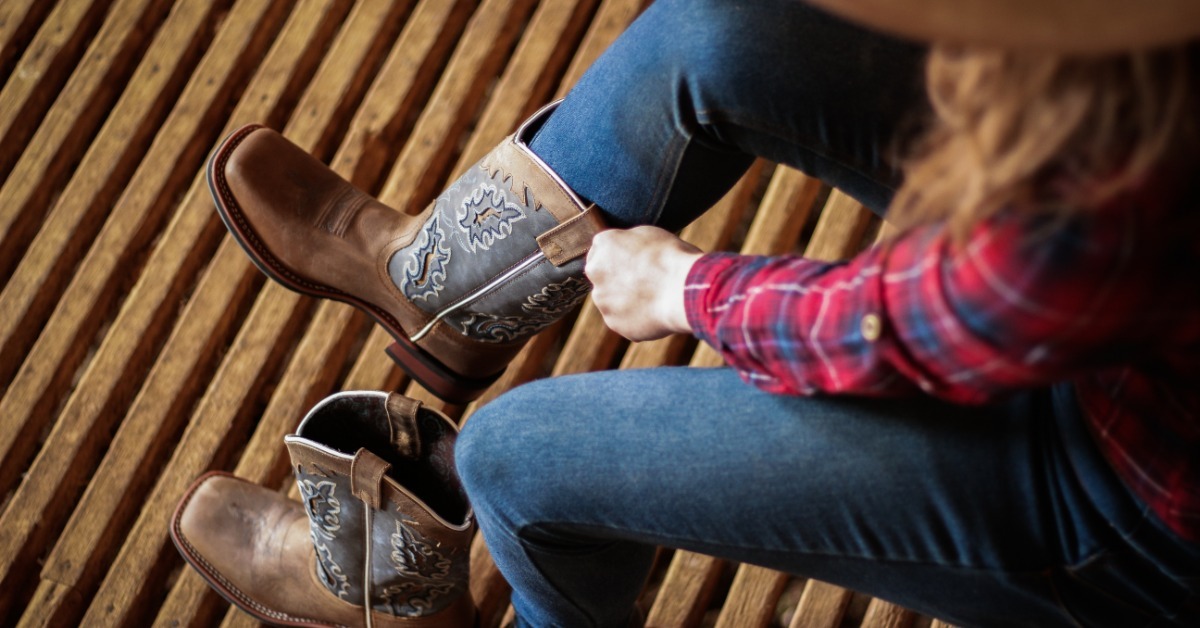 A person wearing blue jeans and a red plaid shirt pulling on a brown western boot beside a matching boot.