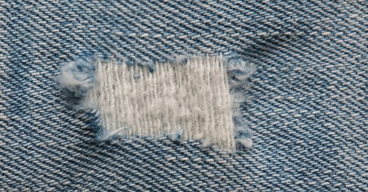 A close-up of light blue denim fabric with a frayed rectangular tear and exposed white threads across the center.