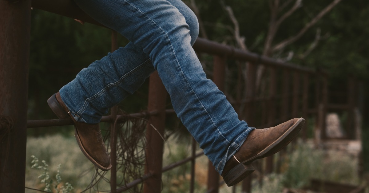 A person wearing blue jeans and brown cowboy boots sitting on a metal fence surrounded by grass and trees.
