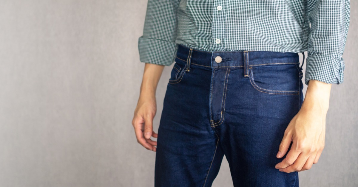 A person wearing a green checked button-up shirt tucked into dark blue jeans against a plain gray wall.