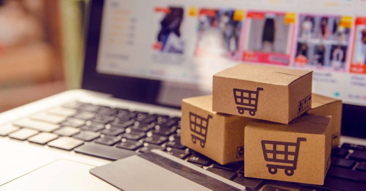Cardboard boxes with shopping cart symbols stacked on a laptop keyboard beside a credit card and screen.