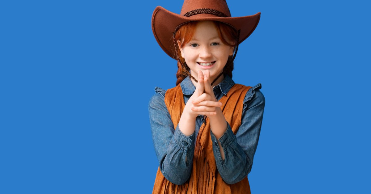 A smiling child wearing a brown cowboy hat, denim shirt, and fringed vest while pointing fingers forward.