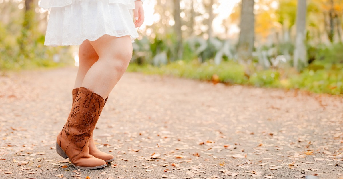 A child in a white dress and brown cowboy boots standing on a leaf-covered path with trees in the background. A child in a white dress and brown cowboy boots standing on a leaf-covered path with trees in the background.