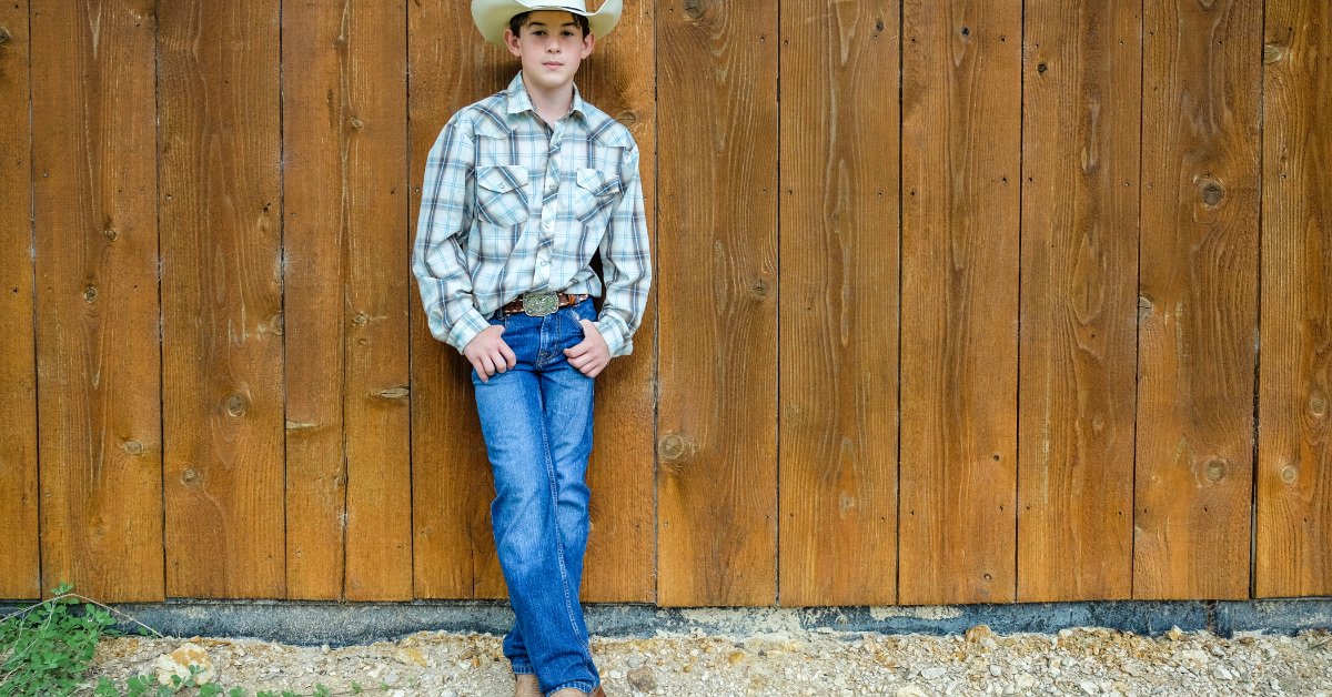 A boy wearing a white cowboy hat, blue plaid shirt, jeans, and boots leaning against a tall wooden barn wall.