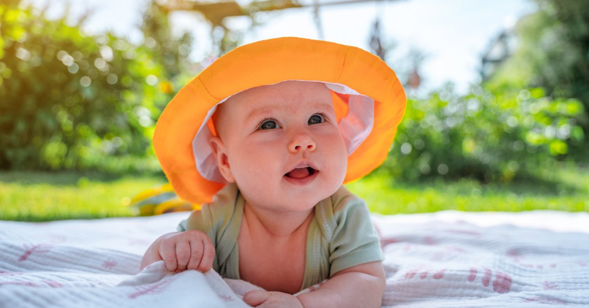A baby wearing a bright orange sun hat and pale green bodysuit lying on their stomach on a blanket outdoors.