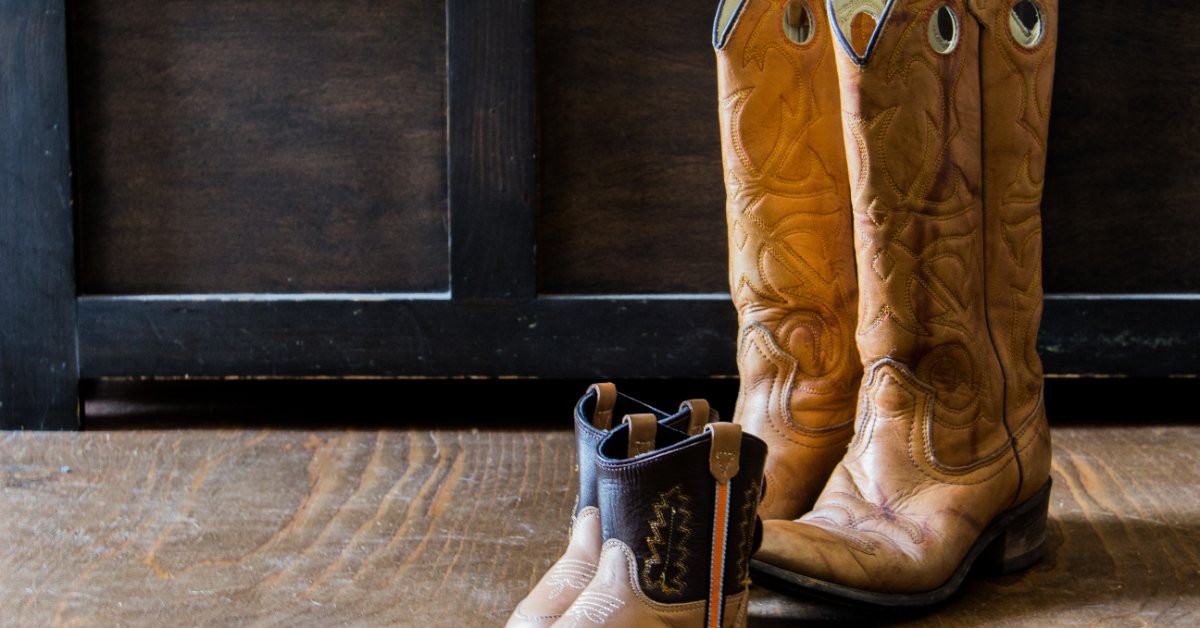 A pair of small brown cowboy boots positioned in front of a pair of tall tan leather cowboy boots on a wooden floor.