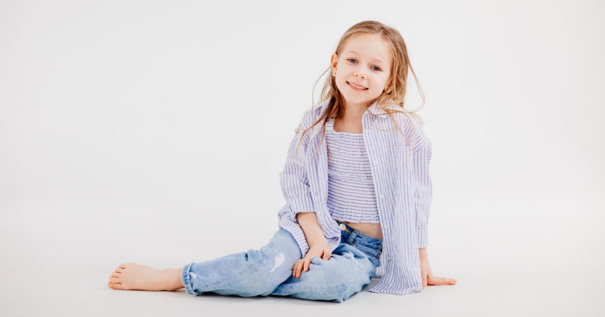 A little girl with blonde hair sitting posed against a white background. She is wearing blue jeans and a striped shirt.