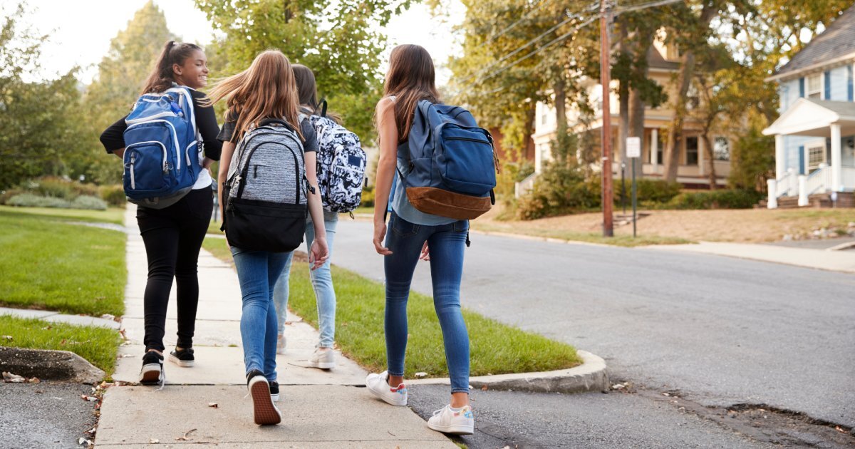 Four young girls walking on a sidewalk in a neighborhood. They're wearing jeans and shirts and carrying backpacks.