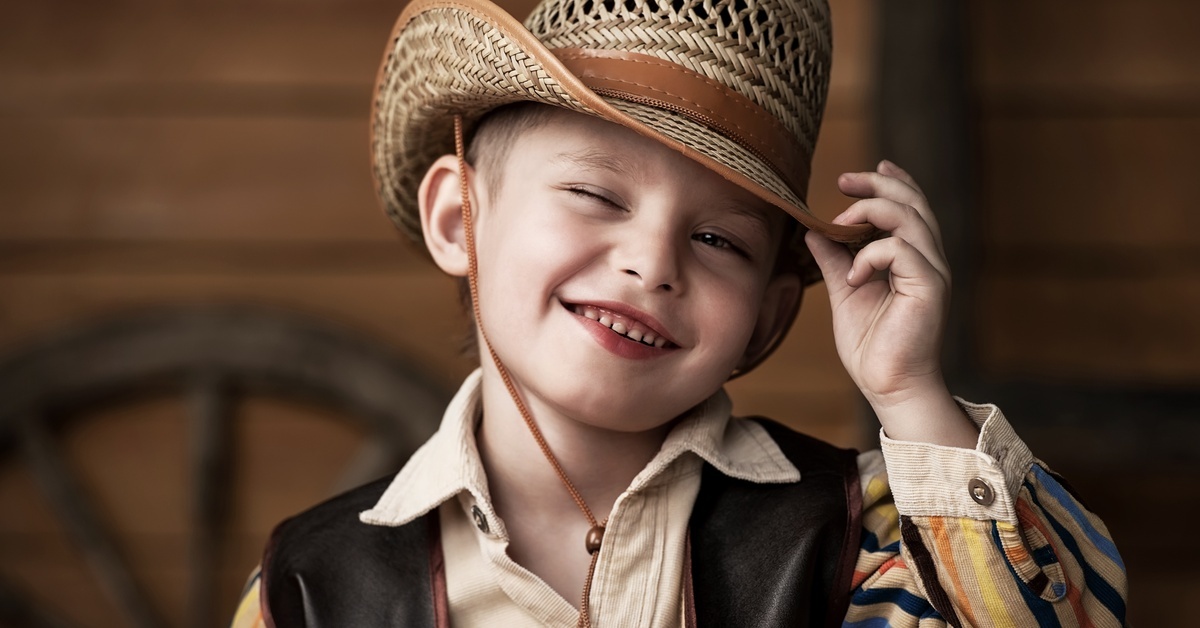 A little boy smiling and tipping his cowboy hat. He is wearing a striped shirt and a leather Western vest.