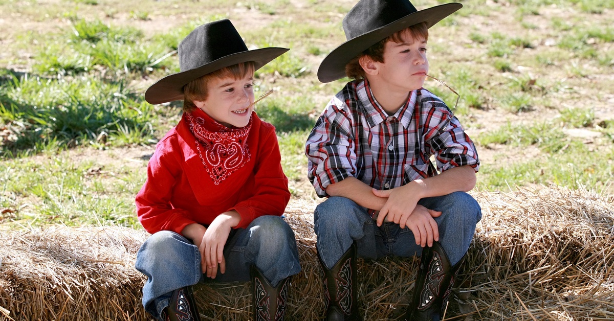 Two boys sitting on hay bales in cowboy boots, jeans, and cowboy hats. One is wearing a red bandana.