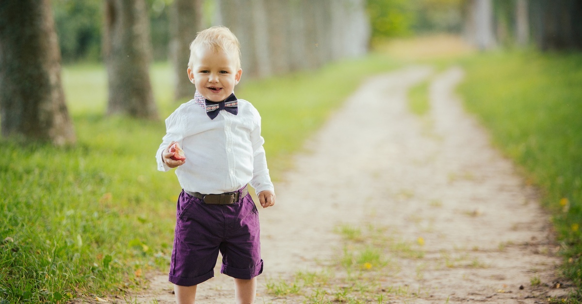 A little boy in purple shorts, a belt, a white button-up, and a bowtie smiling while standing on a grassy dirt road.
