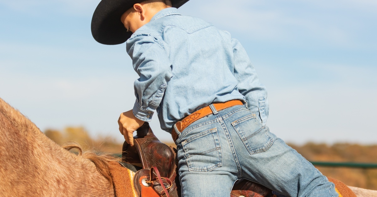 A little boy wearing jeans, a belt, a denim shirt, and a black cowboy hat. He is climbing up into a saddle.