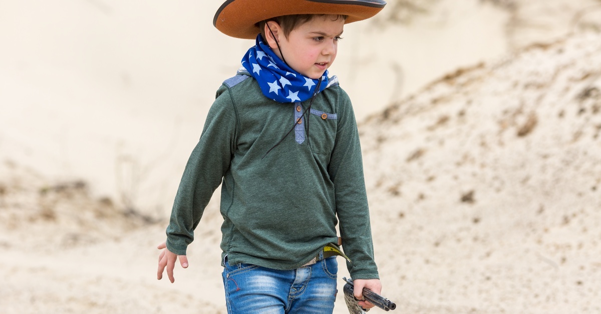 A little boy wearing a bandana and a cowboy hat with a sheriff's badge on the front. He is holding a toy pistol.