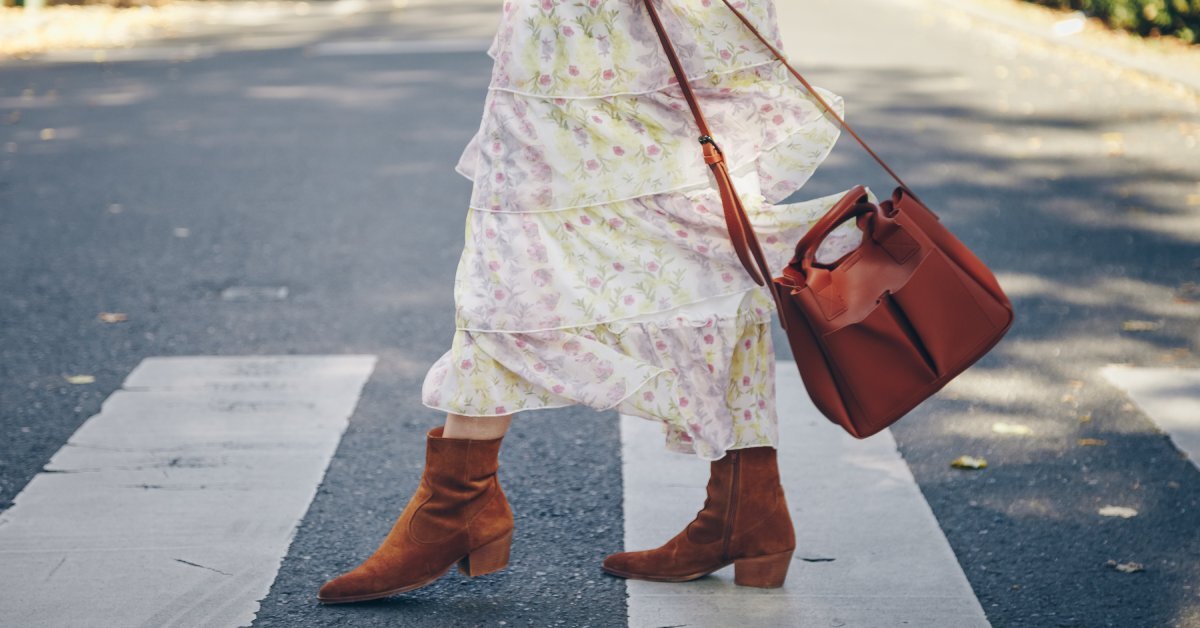 A woman in brown ankle boots and a layered floral skirt walks on a crosswalk. She is carrying a purse.