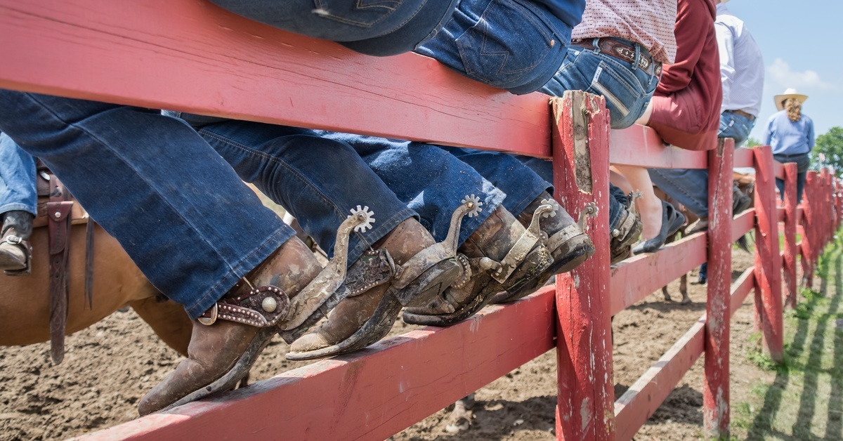 A row of people wearing blue jeans and dirty boots with spurs sitting on a red wooden fence.