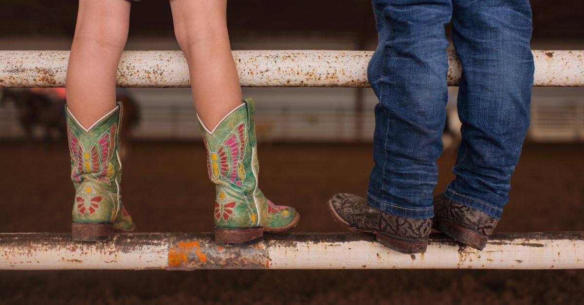 A close-up of two kids wearing cowboy boots while standing together on the round, rusty metal bar of a barn gate.
