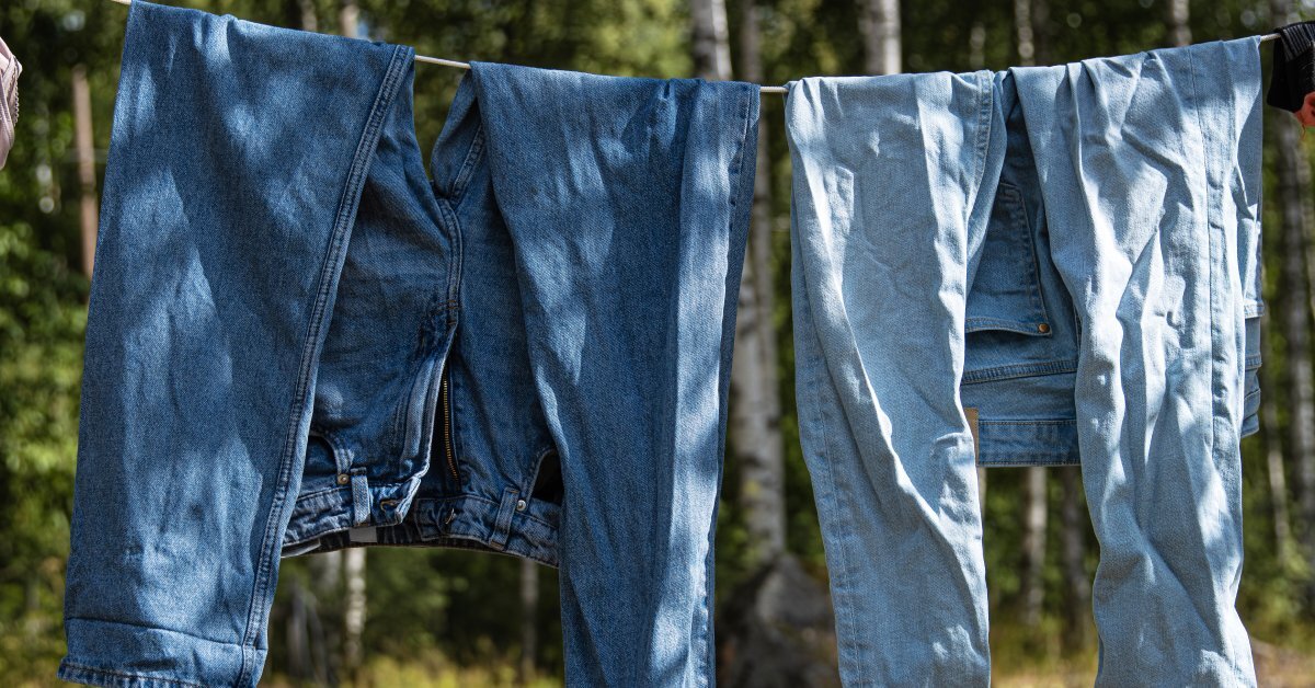 Two pairs of blue jeans hanging on a clothesline outside. There are trees around them and they are partially in shade.