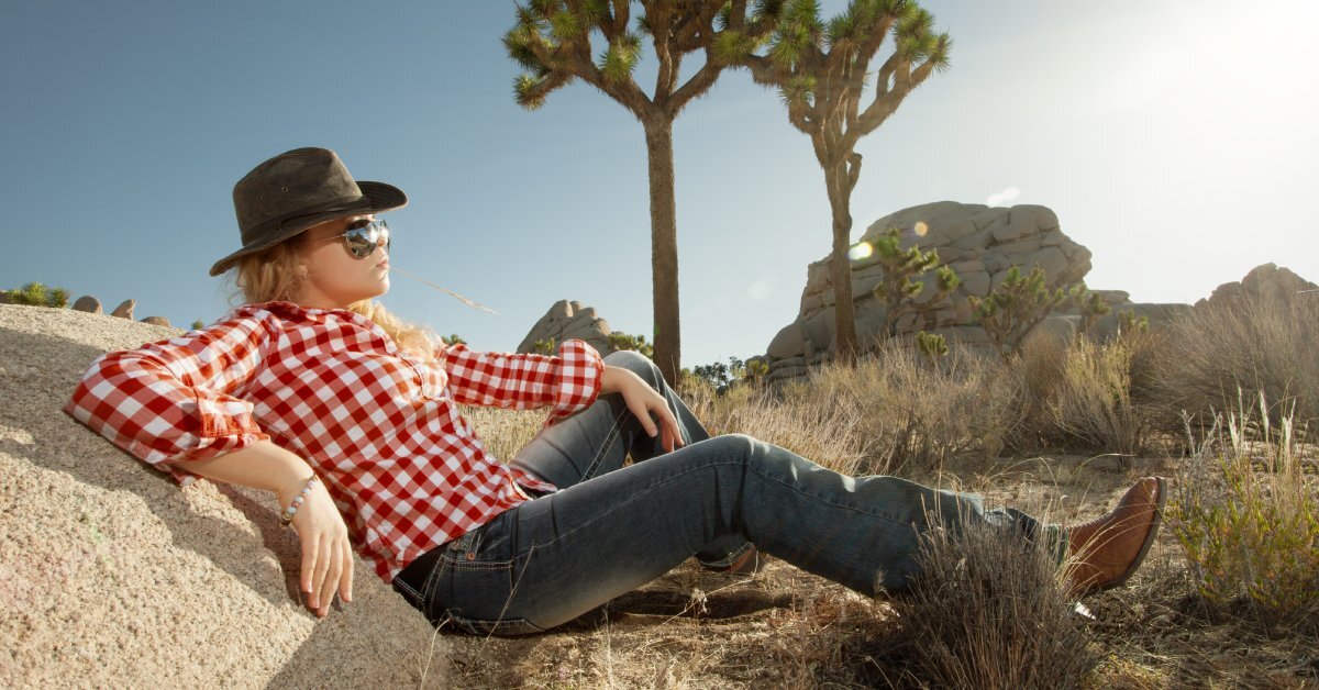 A woman wearing boots, jeans, a checkered shirt, and a cowboy hat sits on the ground beneath the sun.