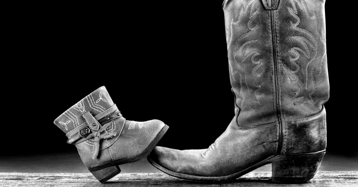 A baby-sized cowboy boot with its toe resting atop the toe of a regular sized cowboy boot.