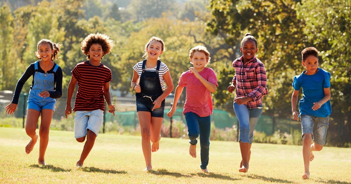 A group of kids running barefoot through a field. They are wearing jeans or overalls and t-shirts.