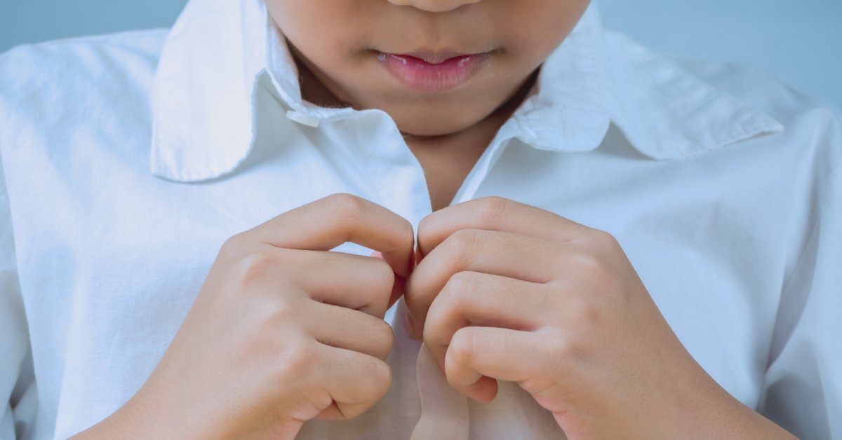 A little kid wearing a white button up shirt. They are looking down and buttoning the top button.