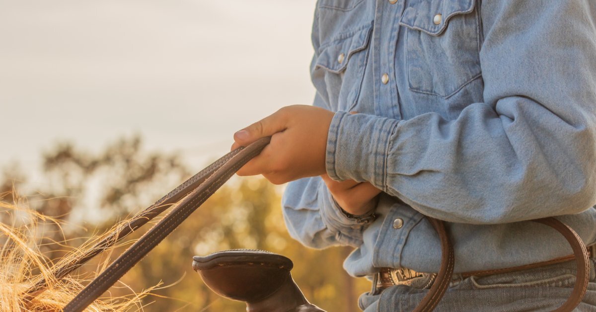 A little kid in a saddle holding the horse's reins. They are wearing a denim shirt with pearl snaps.