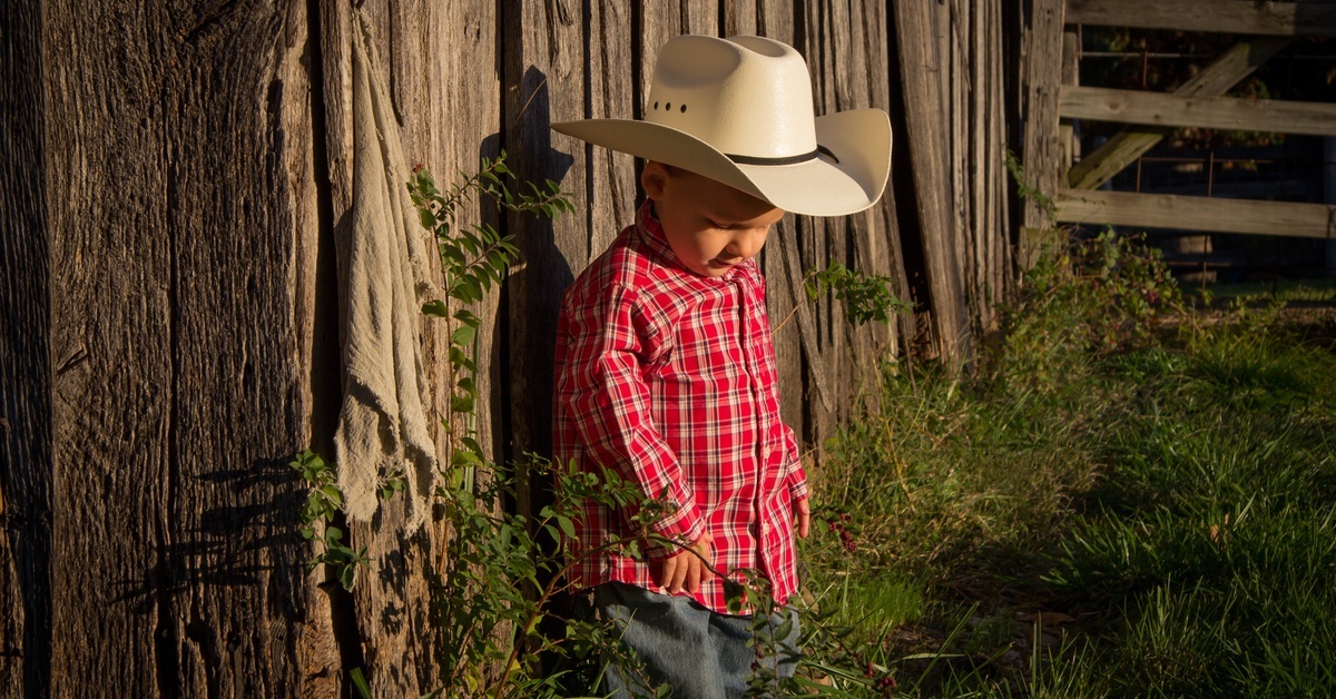 A child in red flannel and a Western hat standing against a wooden fence and looking down as a light casts his shadow.