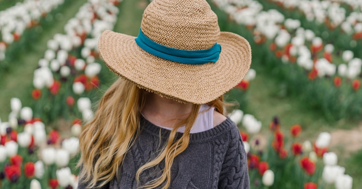 A child with long blonde hair tilting her head forward as her straw hat with a green band covers her face.