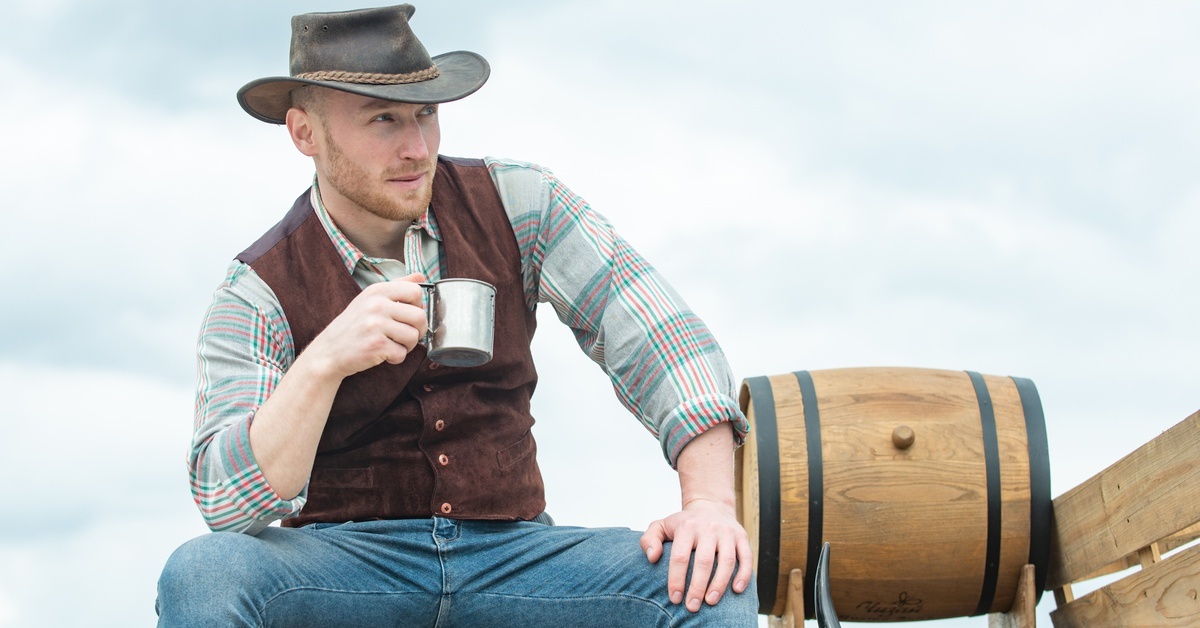 A man in a brown vest, plaid shirt, and a cowboy hat sitting on a bench next to a wooden barrel while holding a mug.