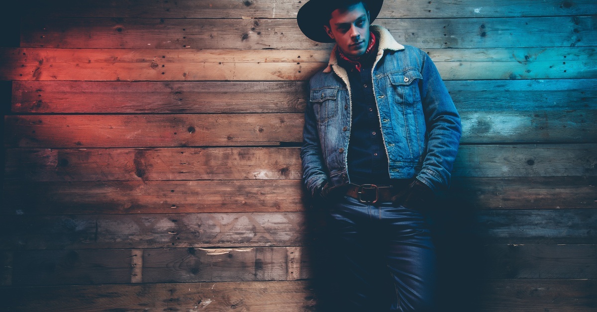 A man in a denim jacket lined with cotton, jeans, and a cowboy hat leaning against a wall in a dimly-lit area.