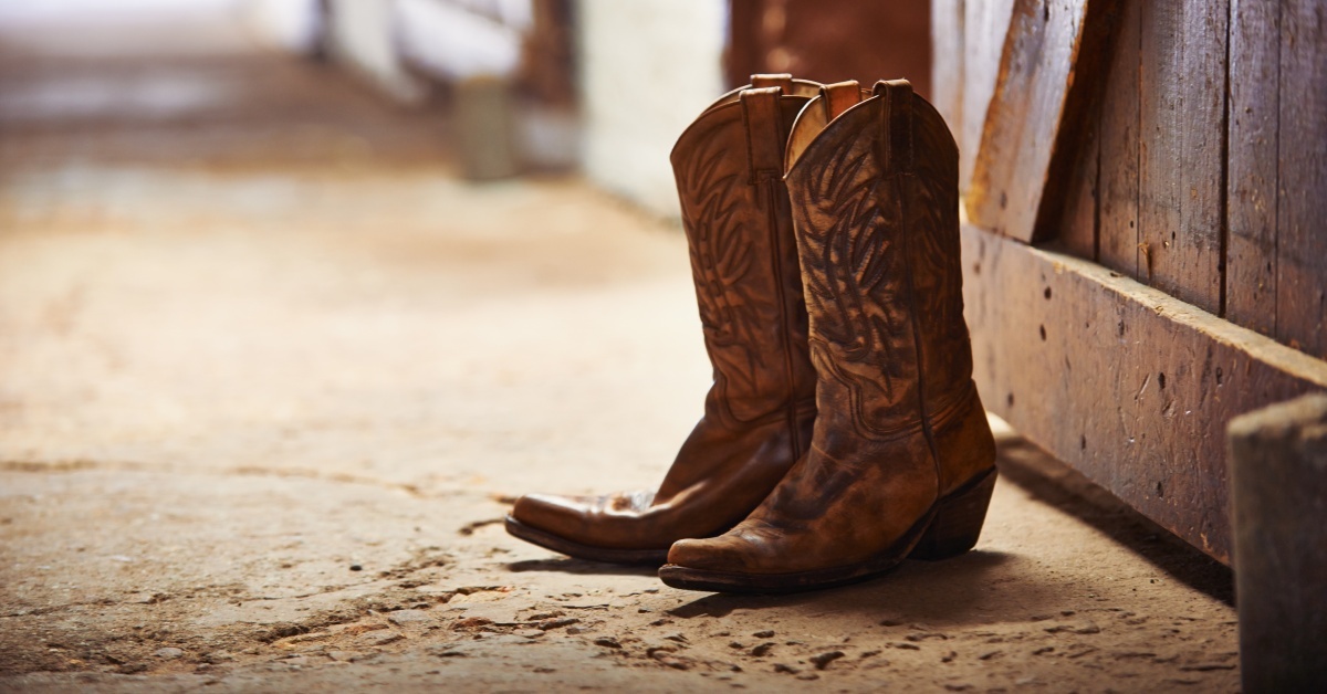 A pair of cowboy boots with multiple impressions and grooves placed by a barn door on a damaged concrete floor.