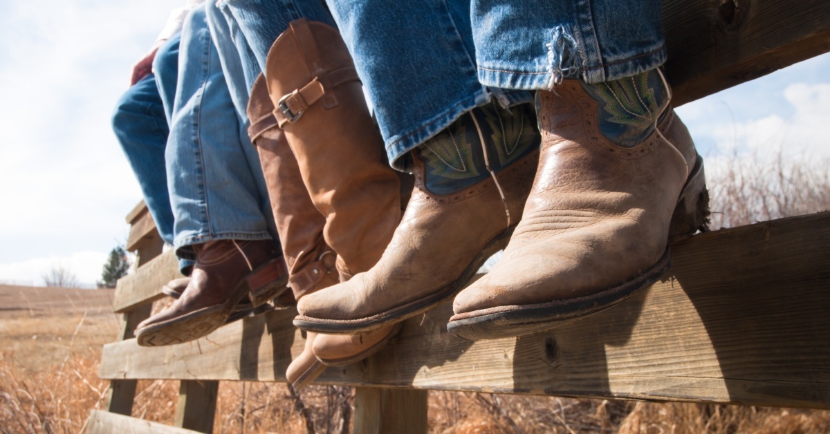 Three people sitting on a wooden fence during the day wearing blue jeans and cowboy boots of different styles.
