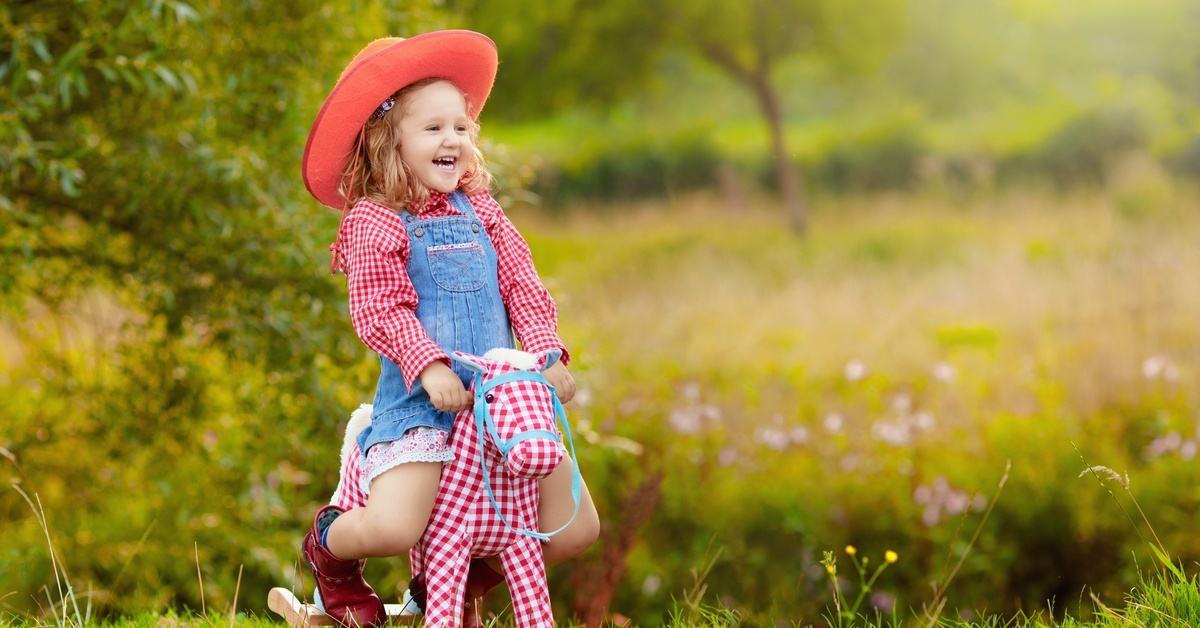 A smiling girl wearing a pink flannel with overalls and red cowgirl boots rides a toy rocking horse outside.