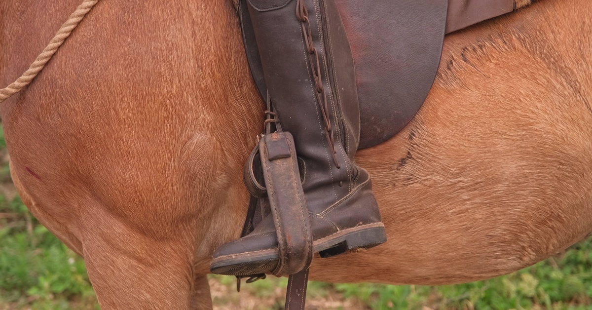 A person wearing long, brown leather cowboy boots sitting on a brown horse with their legs in the stirrups.