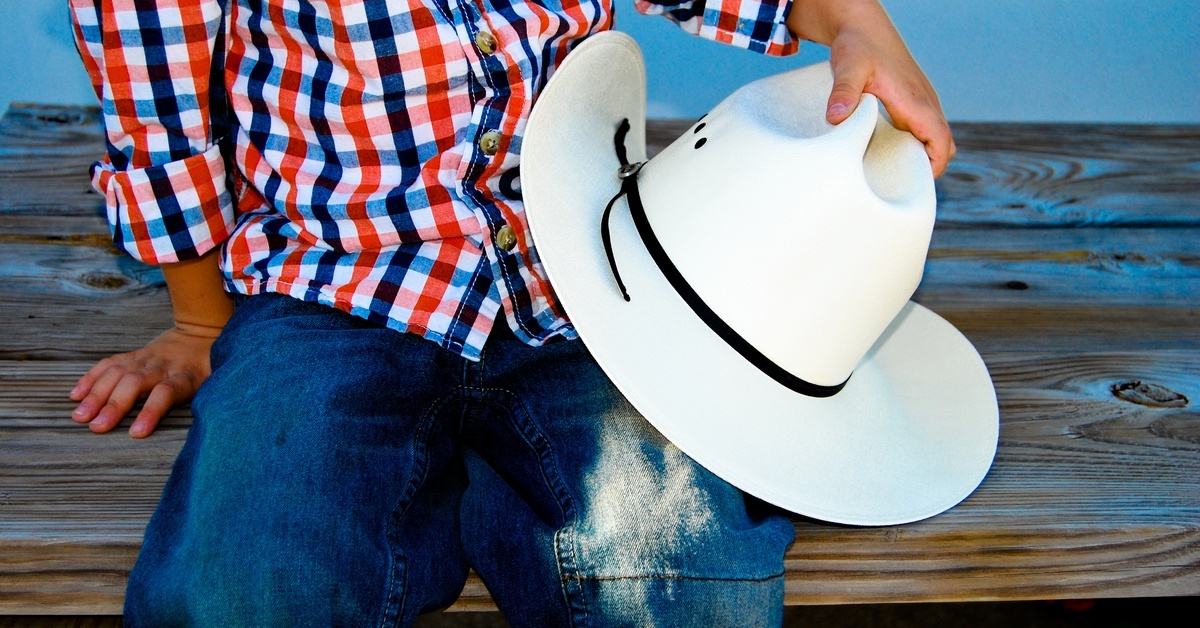 A child wearing a flannel shirt with red white, and blue checks and stained blue jeans while holding a cowboy hat.