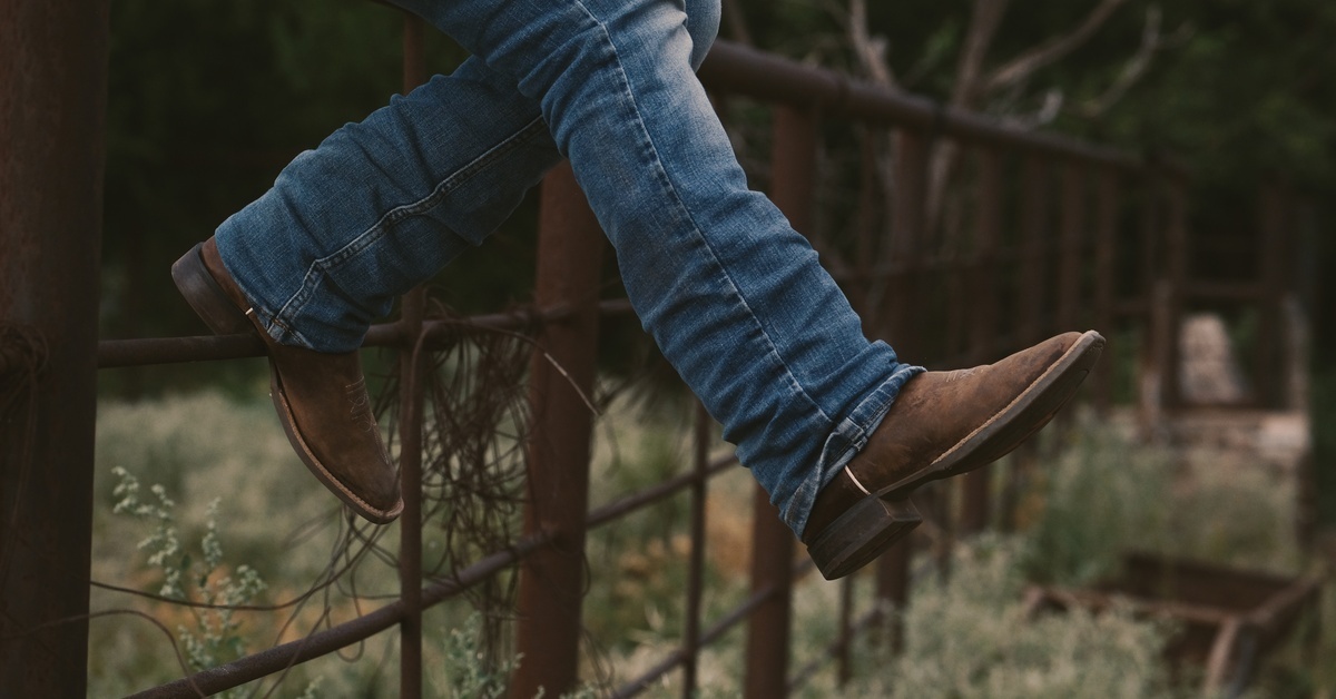 A close-up of a child's jeans-clad legs in cowboy boots, with one foot kicked out as they sit on a wooden fence.