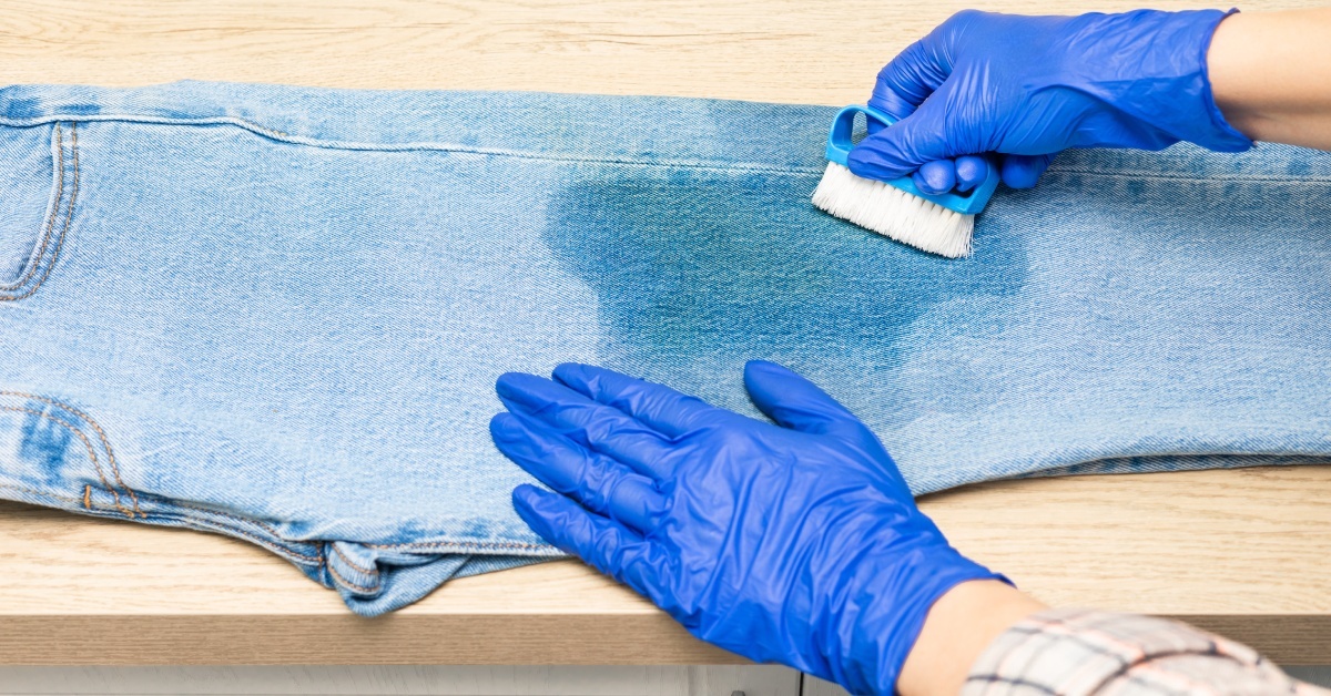 A person wearing blue gloves is trying to remove the stain on a pair of jeans using a small blue brush.