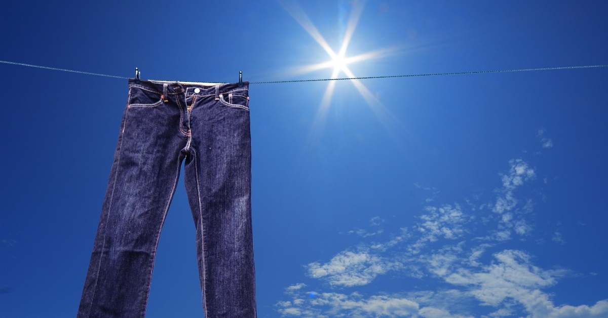 A pair of blue jeans hanging from a clothes line under a lightly-cloudy sky as the sun shines bright above.