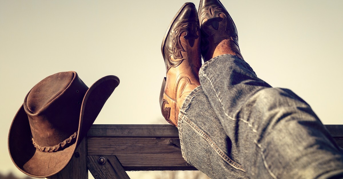 A person wearing jeans and Western boots crosses their ankles atop a fence. A Western hat rests on the edge of the fence.