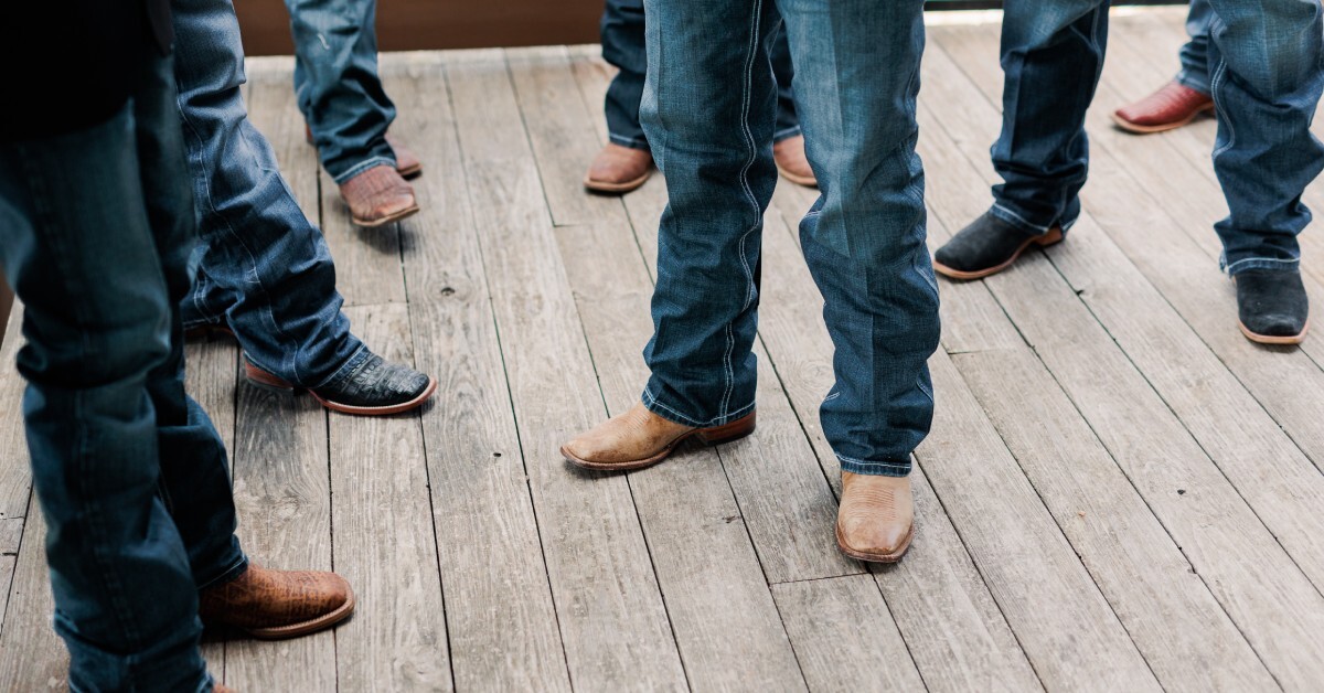 Multiple people are wearing various styles of cowboy boots and relaxed-fit jeans. They stand on a wooden deck.