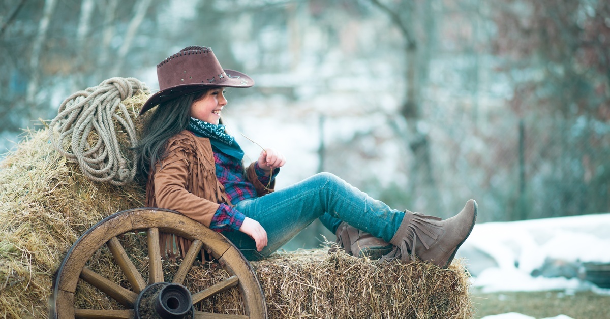 A smiling young girl wearing a cowboy hat, tasseled boots, and other Western attire sitting on a stack of hay bales.