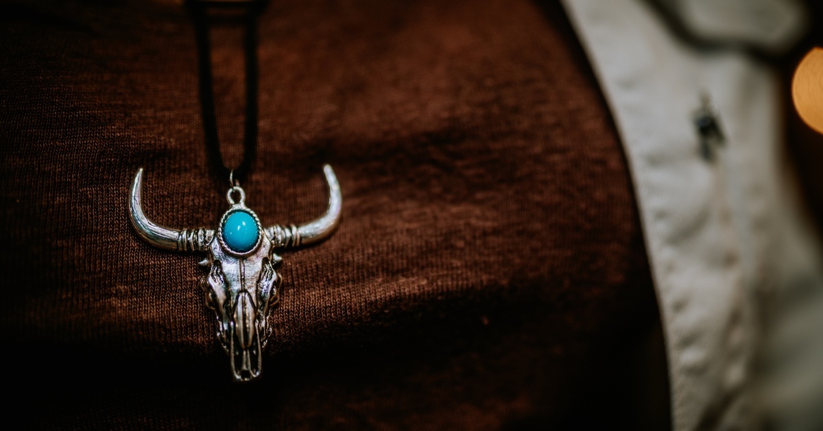 A necklace with a cow skull and a turquoise gem rests on a person wearing a red shirt and pale-green jacket.