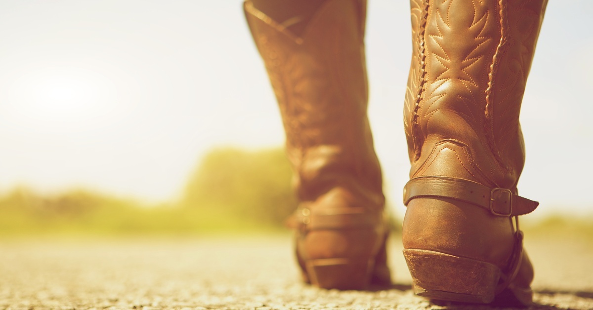 A person walking away on a dirt road, wearing cowboy boots with a low heel, leather straps, and etchings.