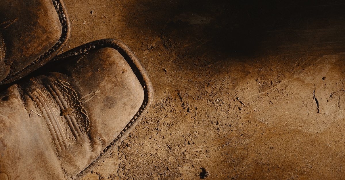 A pair of dirtied brown cowboy boots with a square toe and threaded design, placed on a patch of compacted dirt.
