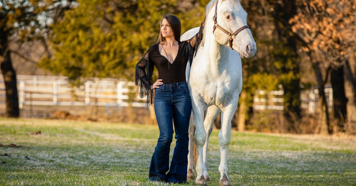 A woman in bootcut jeans and a black shirt with tassels poses and leans on a white horse in a field.